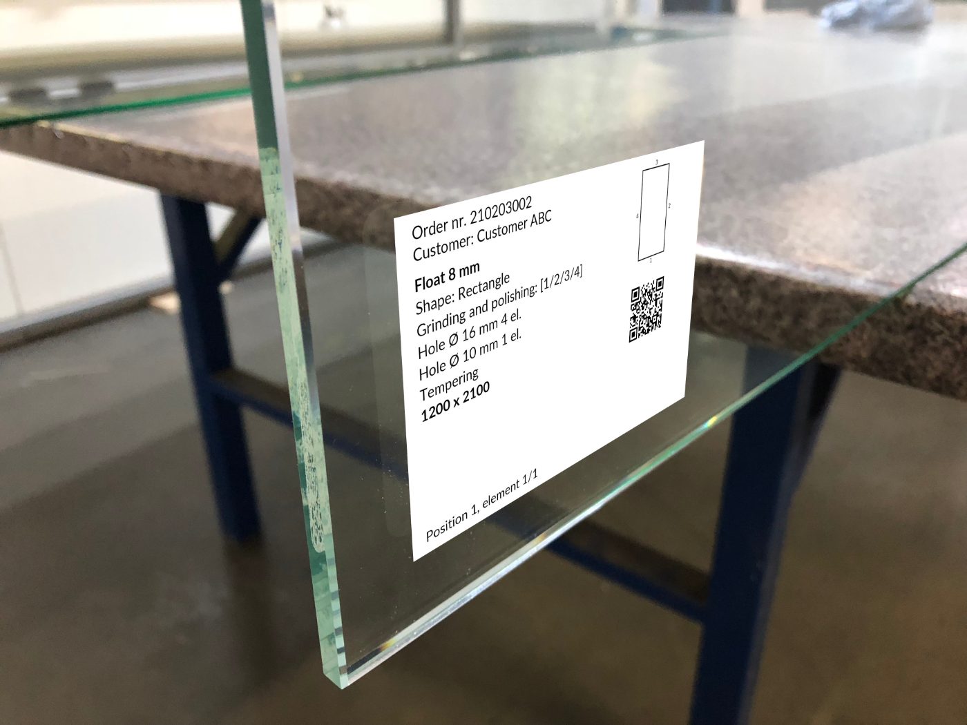 Glass pane on a workshop table with a printed MonitGlass QR label - order details, grinding, holes and tempering